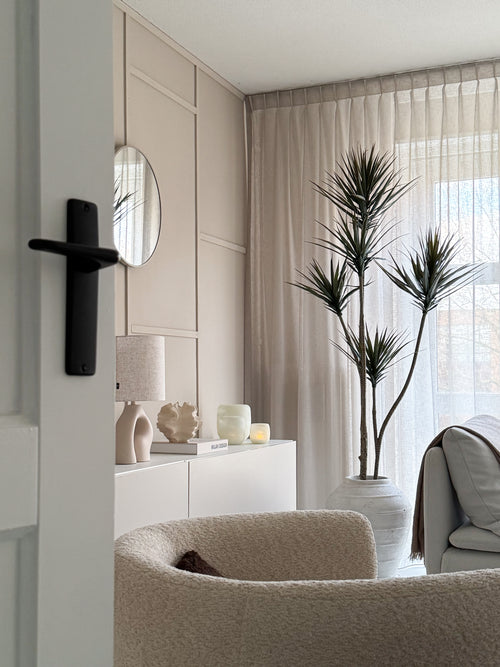 Een moderne, minimalistische woonkamer met een beige fauteuil met textuur, Kunstplant Dracaena Fragnans 180 cm, wit dressoir met decor, ronde spiegel en transparante gordijnen die zacht daglicht filteren door het raam.