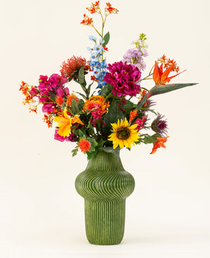 Een groene vaas met structuur toont het Zijden kunstbloemen boeket Colourful Spirit, met zonnebloemen, roze pioenrozen, oranje en paarse bloemen en weelderig groen tegen een effen lichte achtergrond.