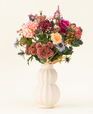 In een witte, gebogen vaas van keramiek staat het Zijden kunstbloemen boeket Pretty Pastel - een kleurrijke mix van duurzame bloemen zoals roze rozen, dahlia's, paarse en oranje bloemen en groen - prachtig gerangschikt tegen een lichte achtergrond.