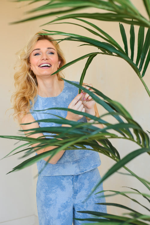 Een glimlachende vrouw met lang blond haar, in een lichtblauwe mouwloze top en bijpassende broek, staat binnen achter de Kunstplant Kentia Palm 180 cm en raakt speels een van de groene bladeren aan.