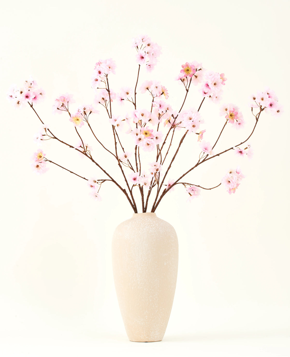 Het Zijden kunstbloemen boeket Kersenbloesem roze 114 cm heeft lange, dunne takken met trossen lichtroze bloemen in een beige vaas van keramiek, die een opvallend contrast vormen tegen de effen witte achtergrond.
