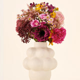 Een boeket van Zijden kunstbloemen, A Touch of Ashley, in roze, paarse en gele tinten, gepresenteerd in een off-white vaas met bubbelvorm en een gestructureerd oppervlak.