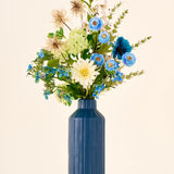 Een boeket van blauwe, witte en groene Zijden kunstbloemen, het boeket Blue Whisper, in een hoge, geribbelde blauwe vaas.