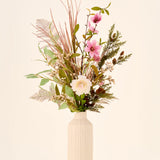 Zijden kunstbloemenboeket Frosted Blush met kunstbloemen in roze, witte bloesems, grassen en groen in een geribbelde beige vaas.