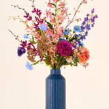 Een groot boeket kleurrijke kunstbloemen van Zijden, het Mikaya-boeket, met onder meer lelies, dahlia’s en orchideeën, in een hoge, geribbelde blauwe vaas.