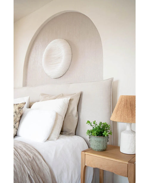 Een gezellige slaapkamer met een beige hoofdeinde, neutraal beddengoed en kussens, een houten nachtkastje met daarop de Kunstplant Eucalyptus 27 cm, een lamp met een getextureerde kap en ronde witte muurkunst boven het bed in een gewelfde nis.