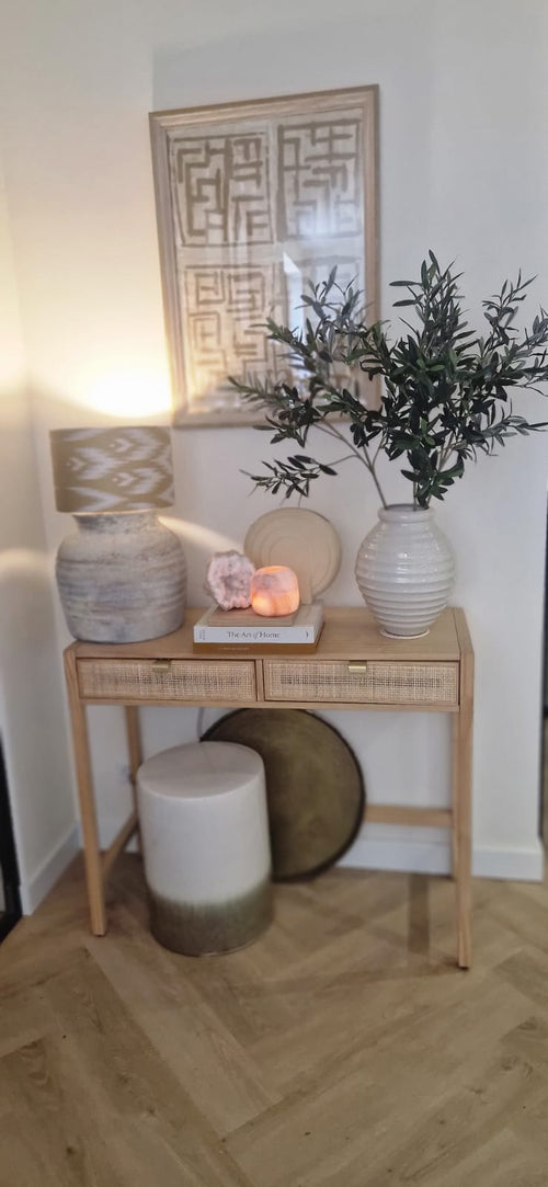 Een houten consoletafel met twee laden bevat een lamp, een grote witte vaas met kunst olijfblad 90 cm brandvertragend, een kleinere vaas, boeken en decoratieve stenen. Mediterrane accenten en ingelijste abstracte kunst maken de look hierboven compleet. Onderaan staan een kruk en een rond dienblad.
