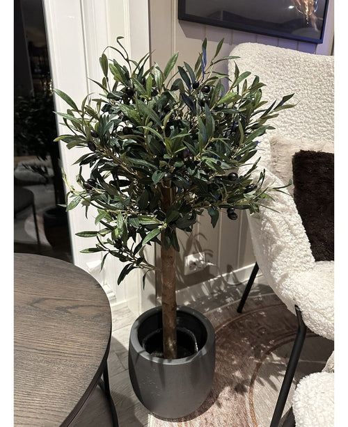 De Kunst olijfboom UV 85 cm, een compacte en onderhoudsvriendelijke kunstplant met donkergroene bladeren en zwarte olijven, staat binnen naast een ronde houten tafel en een fauteuil met witte textuur.