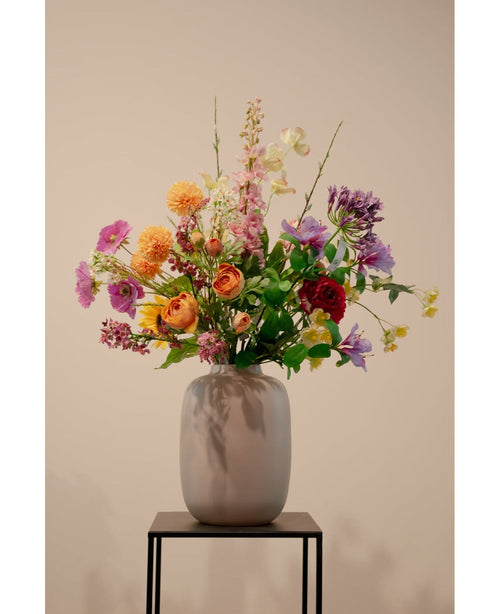 Een Kunstkornoelje Real Touch Lichtgeel 74cm kunstbloemstuk zit in een keramieken vaas op een zwart metalen standaard, tegen een effen beige achtergrond.