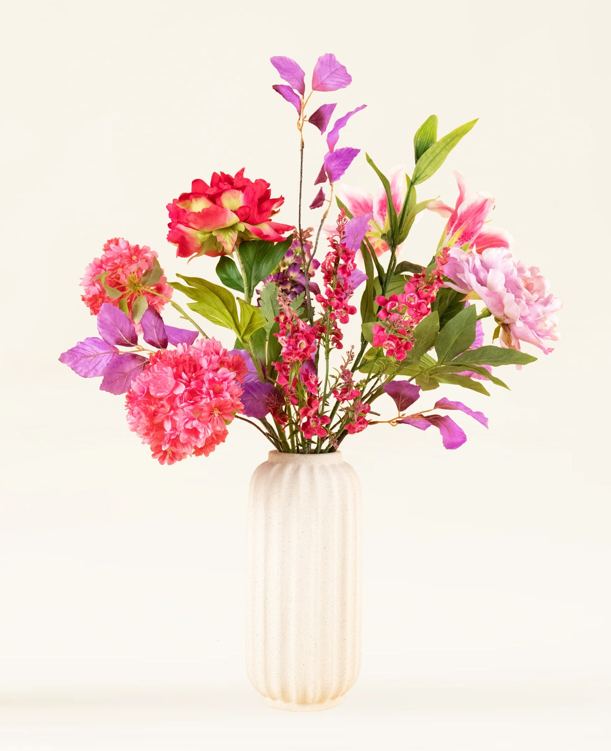 Een crèmekleurige, geribbelde vaas van keramiek bevat het Zijden kunstbloemen boeket Rosy Saga Mist, met roze, rode en paarse zijden bloemen met groene bladeren tegen een lichte achtergrond.