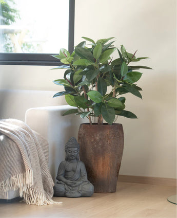 Een Kunstplant Rubber deluxe 90 cm staat op een lichte houten vloer naast een grijs Boeddhabeeld, met zonlicht dat op een nabijgelegen bank valt die is gedrapeerd in een beige en witte deken met franjes.