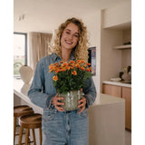Een vrouw met krullend blond haar, gekleed in spijkerkleding, glimlacht terwijl ze de Kunstplant Daisy UV 31 cm – een pot met feloranje en gele bloemen – vasthoudt in een moderne keuken.