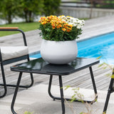 Een zwarte terrastafel met een Kunstplant Daisy UV 31 cm – een ronde witte pot met levendige oranje en witte madeliefjes – staat naast een zwembad, omringd door tuinstoelen en met groen op de achtergrond.