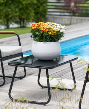 Een zwarte terrastafel met een Kunstplant Daisy UV 31 cm – een ronde witte pot met levendige oranje en witte madeliefjes – staat naast een zwembad, omringd door tuinstoelen en met groen op de achtergrond.