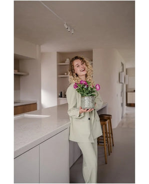 Een glimlachende vrouw met krullend haar, gekleed in een lichtgekleurd pak, staat in een moderne, minimalistische keuken met in haar handen een Kunstbloem Petunia UV 25 cm donkerroze met levendig roze bloemen.
