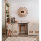 Een jong meisje in een lichte jurk staat naast een plank waarop de Kunst-hangplant ‘Frosted Varen op bal 76 cm’ en andere hangplanten staan, in een moderne kamer met beige kasten, een hoge kruk, een wandlamp van riet en een geruit vloerkleed op de vloer.