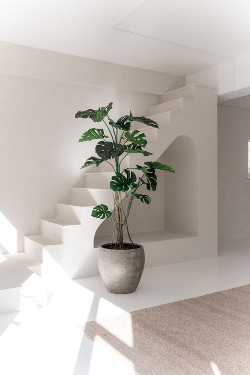 De Kunstplant Monstera 210 cm, met brede groene bladeren, staat op een witte vloer in een lichte, minimalistische kamer met witte muren, een trap en natuurlijk zonlicht.