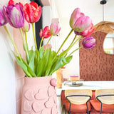 Een roze vaas met hartjes in reliëf houdt de Kunstbloem Franse Tulp 60 cm rood op een eettafel. Daarachter staan stoelen met rieten ruggen, een taart op tafel en een spiegel op een gedessineerde bruine muur onder een geweven hanglamp.