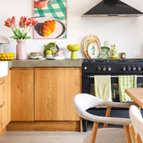 Een lichte keuken met houten kasten, een zwart fornuis, groene handdoeken, kleurrijk servies en een ingelijste afbeelding van een gebakje op een geruite tafel krijgt een frisse uitstraling dankzij de Kunstbloem Franse Parkiet Tulp 76 cm rood/geel. Een gezellige witte stoel staat naast een licht houten eettafel.