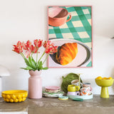 Een aanrecht met allerlei kleurrijke serviesstukken en een Kunstbloem Franse Parkiet Tulp van 76 cm in rood/geel in een roze vaas. Daarboven hangt een ingelijste afbeelding van een croissant en een theekopje op een groen geruit tafelkleed.