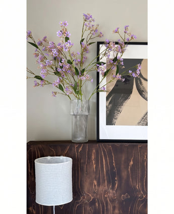 In een doorzichtige vaas staan de bloemen van de Kunsttak Daphne Spray 108 cm paars op een donkerhouten oppervlak naast een modern, abstract kunstwerk in een lijst, met daaronder een witte lampenkap met structuur.