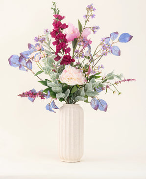 Een vaas met witte structuur toont het "Zijden kunstbloemen boeket Amethyst Romance" met roze pioenrozen, paarse en blauwe bloemen, bordeauxrode bloemen en groen gebladerte tegen een lichte, neutrale achtergrond.