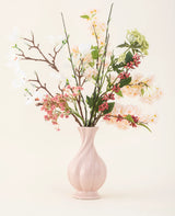 Een roze vaas van keramiek bevat het Zijden kunstbloemen boeket Springtime Poem, met witte, roze en groene bloemen met groene bladeren en bruine stelen, tegen een lichte achtergrond.