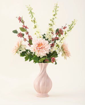 Een roze vaas van keramiek bevat het Zijden kunstbloemen boeket Morning Whisper, met lichtroze dahlia's, witte leeuwenbekjes, groene bladeren en trosjes kleine rode besjes, tegen een effen gebroken witte achtergrond.