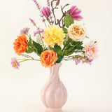 Het Zijden kunstbloemen boeket Sweet Daybreak heeft een pastelroze vaas gevuld met oranje rozen, gele dahlia's, roze magnolia's en lichtpaarse bloesems, prachtig gerangschikt tegen een effen lichte achtergrond.