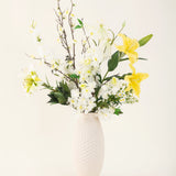 Een vaas met witte structuur toont de elegantie van het Zijden kunstbloemen boeket Sunkissed Ivory, met gele lelies, witte bloemen en groene bladeren tegen een effen lichte achtergrond.