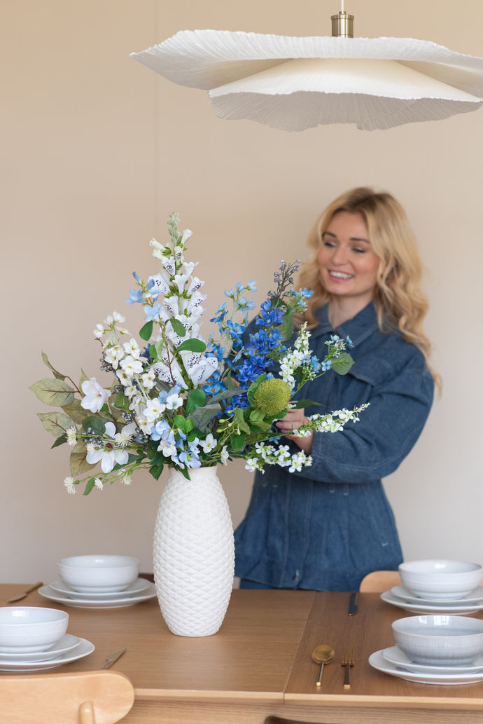 Een glimlachende vrouw in een blauwe spijkerjurk schikt het Zijden kunstbloemen boeket Cool Breeze in een gestructureerde witte vaas op een eettafel gedekt met wit servies en goud bestek.