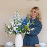 Een glimlachende vrouw in een blauwe spijkerjurk schikt het Zijden kunstbloemen boeket Cool Breeze in een gestructureerde witte vaas op een eettafel gedekt met wit servies en goud bestek.
