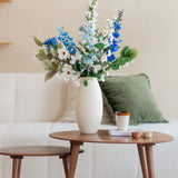 Een Zijden kunstbloemen boeket Cool Breeze van blauwe en witte bloemen staat in een witte vaas op een ronde houten salontafel met een kopje, kaarsen en een boek. Daarachter staat een witte bank met groene en beige kussens.