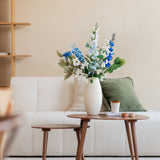 Een moderne woonkamer met een witte bank, een groen kussen en een klein houten tafeltje met daarop een Zijden kunstbloemen boeket Cool Breeze in een witte vaas, plus een koffiekopje en boeken, tegen een beige muur met een minimalistische plank.