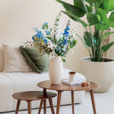 In een gezellige woonkamer staan twee houten nesttafels met een witte vaas met daarin het Zijden kunstbloemen boeket Cool Breeze, plus een kopje, boek en kaars. Een witte bank met groene kussens en een grote plant in pot maken het luchtige decor compleet.