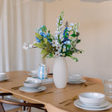 Een houten eettafel gedekt met borden, schalen, goudkleurig keukengerei en een witte vaas gevuld met het Zijden kunstbloemen boeket Cool Breeze. Zachte verlichting en moderne witte hanglampen erboven creëren een uitnodigende sfeer.