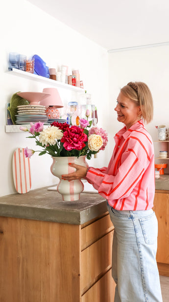 Een vrouw in een roze gestreept shirt schikt het luxe bloemen cadeau, Zijden kunstbloemen boeket Dream Delight, in een gestreepte vaas op het aanrecht, met achter haar planken met borden en glazen zichtbaar.