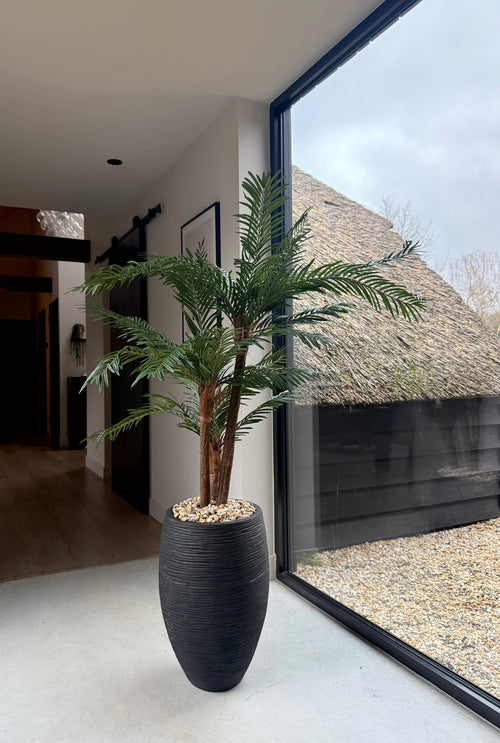 De Kunstpalm Cycas Classic 135 cm in een strakke sierpot staat bij een groot raam met daglicht. Buiten zijn een rieten dak en grind te zien. Het interieur is modern en minimalistisch.