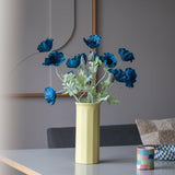Een Kunstbloem Poppy van 73 cm in donkerblauw staat in een gele vaas op een tafel, vergezeld van een klein, kleurrijk gestreept kopje, in een moderne, zacht verlichte kamer met een minimalistische inrichting en neutrale tinten.