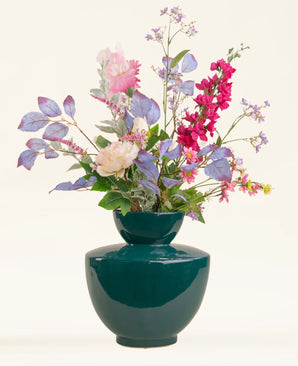 In een glanzende donkerblauwe vaas staat het Zijden kunstbloemen boeket Amethyst Romance, met lichtpaarse bladeren, roze pioenrozen, rode leeuwenbekken en kleine lavendelbloesems tegen een effen lichte achtergrond.