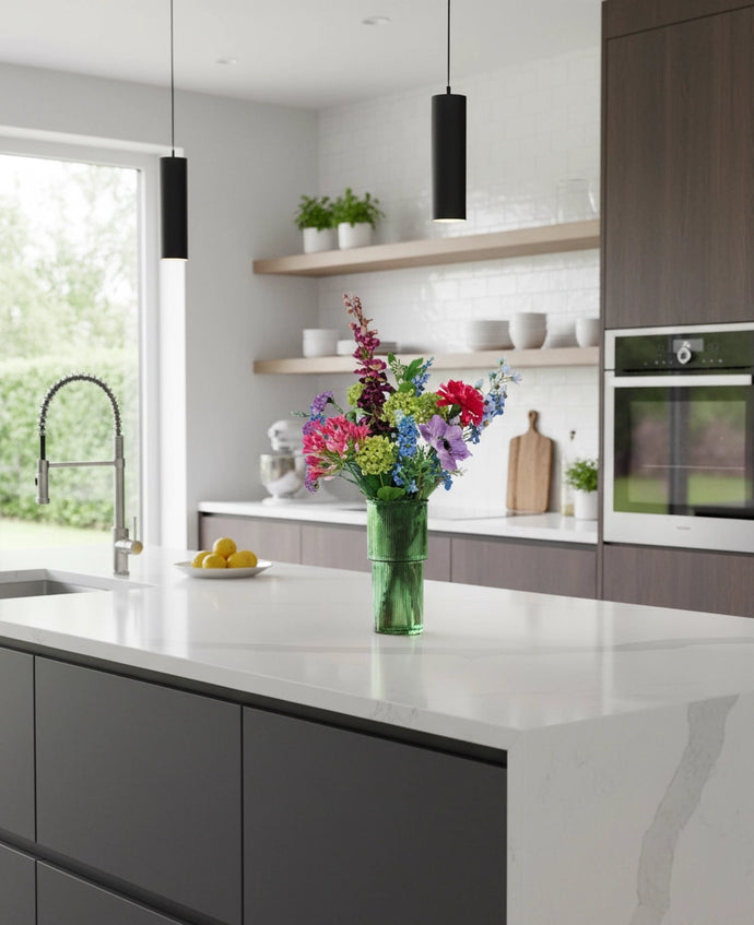Een moderne keuken met donkere kastjes, een wit aanrechtblad en een vaas van het Zijden kunstbloemen boeket Blossom Breeze. Erboven hangen zwarte hanglampen, op open planken staan borden, een schaal is gevuld met citroenen en een ingebouwde oven maakt het plaatje compleet.