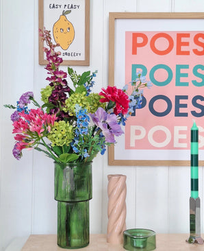 Een groene glazen vaas houdt een Zijden kunstbloemen boeket Blossom Breeze op tafel. Daarachter hangen ingelijste prenten - een met een citroen en "EASY PEASY", een andere met "POESY" - die voor extra charme zorgen. Vlakbij staan een groene kaars en een roze spiraalkaars van Blossom Breeze.