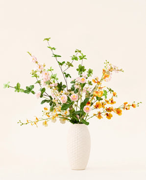 In een vaas met witte structuur staat het Zijden kunstbloemen boeket Blossom Harmony met roze, witte en gele kunstbloemen en groene bladeren tegen een effen lichte achtergrond.