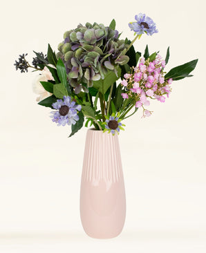 Het Zijden kunstbloemen boeket Blue Belle bestaat uit hortensia's en Blue Belle in paarse, roze en witte tinten, stijlvol geschikt in een hoge lichtroze keramieken vaas tegen een lichte achtergrond.