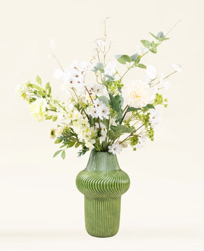 Het Zijden kunstbloemen boeket Claire de Lune heeft witte en lichtgele bloemen met groene bladeren en takken in een groene geribbelde vaas, prachtig tegen een lichtbeige achtergrond.
