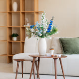 Een moderne woonkamer met een bank met witte structuur, een groen kussen, houten salontafels en een witte vaas met het Zijden kunstbloemen boeket Cool Breeze. Een houten plank met minimale decoratie maakt deze serene omgeving compleet.
