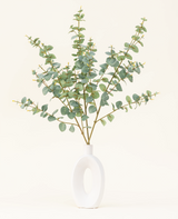 Een moderne witte vaas met een open cirkelvormig ontwerp bevat het Zijden kunstbloemen boeket Eucalyptus, met kunstmatige eucalyptustakken met ronde groene bladeren tegen een effen gebroken witte achtergrond.