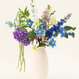 Een vaas met witte structuur bevat het Zijden kunstbloemen boeket Midnight Valley, met levendige blauwe, paarse en gele Real Touch bloemen met verschillende groene bladeren tegen een lichte achtergrond.