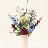 Een witte vaas met het Zijden kunstbloemen boeket Purple Rain toont levendige paarse en witte kunstbloemen en groen blad, een toonbeeld van Nederlands vakmanschap tegen een lichte achtergrond.