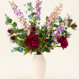 In een vaas met witte structuur staat het Zijden kunstbloemen boeket Moonlit Meadow - rode pioenrozen, roze en paarse riddersporen, blauwe bloemen, groene bladeren en distelachtige bloemen - tegen een effen, lichte achtergrond.